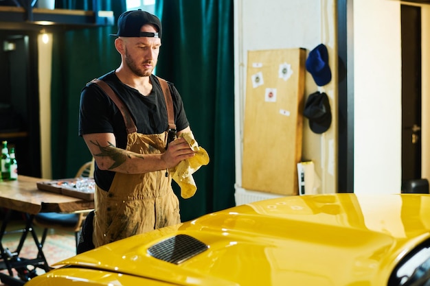 Junger Handwerker in Arbeitskleidung reibt schmutzige Hände mit einem Staubtuch über der Motorhaube