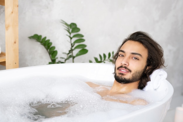 Junger heiterer bärtiger Mann, der sich entspannt, während er im Bad mit heißem Wasser und Schaum vor Kamera liegt