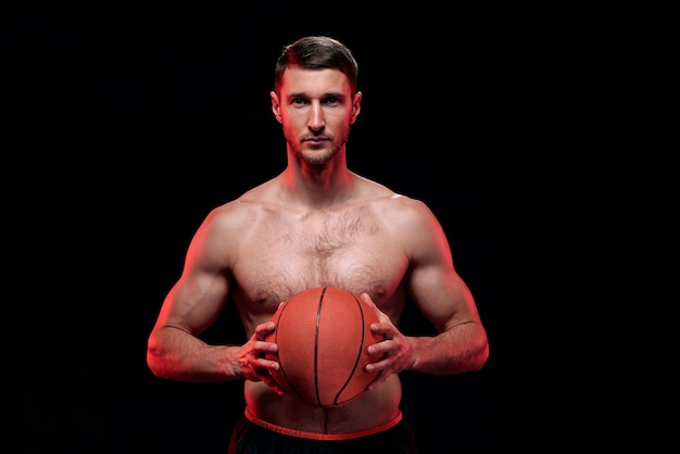Junger hemdloser muskulöser Basketballspieler, der Ball an seiner Brust hält, während er steht und Sie ansieht
