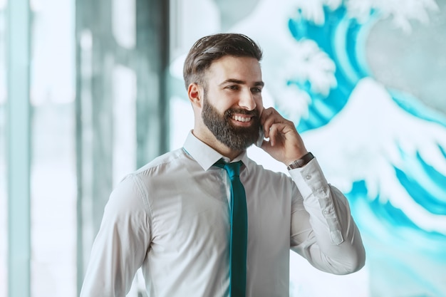 Junger herrlicher lächelnder kaukasischer Geschäftsmann in der formellen Abnutzung, die am Telefon spricht, während er im Büro steht.