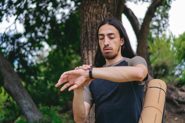 Junger Hippie-Latin-Mann mit langen Haaren, der die Zeit sieht, während er eine Yogamatte in einem Waldgebiet trägt