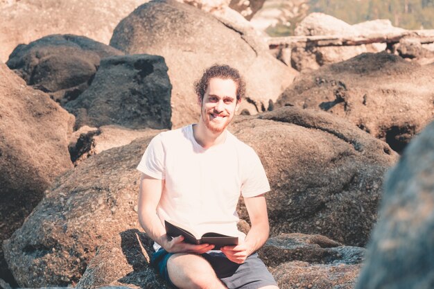 Junger Hippie-Mann, der auf einem weißen Hemd in die Kamera lächelt und ein Buch im Felsen des Strandes liest, während eines Sommertages, Feiertagskälte und Freiheit