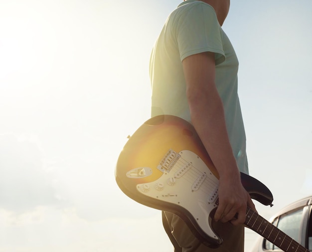 Junger Hipster in blauem T-Shirt mit Gitarre steht im Kontrast zu Sonnenlicht und Blendung auf der Straße