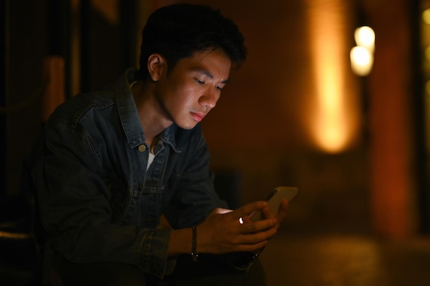 Junger Hipster-Mann mit Handy, der mit Freunden in sozialen Netzwerken chattet, während er abends auf den Straßen der Stadt sitzt