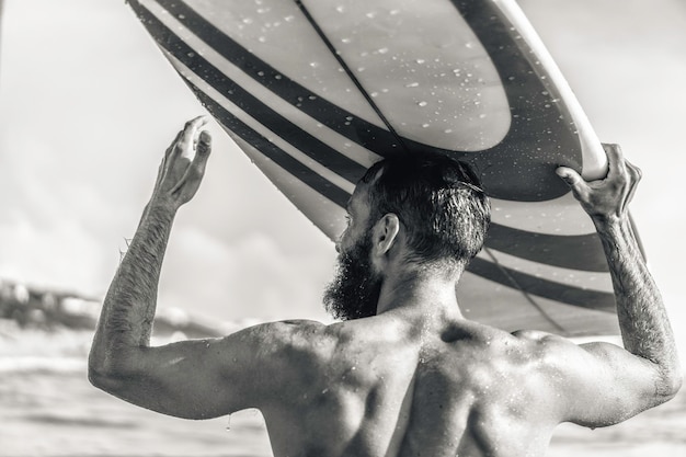 Junger Hipster-Mann mit Surfbrett auf dem Kopf grüßt Freunde mit einer Geste Guy hat Spaß beim Extremsport Sportlicher bärtiger Mann trainiert mit Surfbrett auf dem Meer Schwarz-Weiß-Bearbeitung