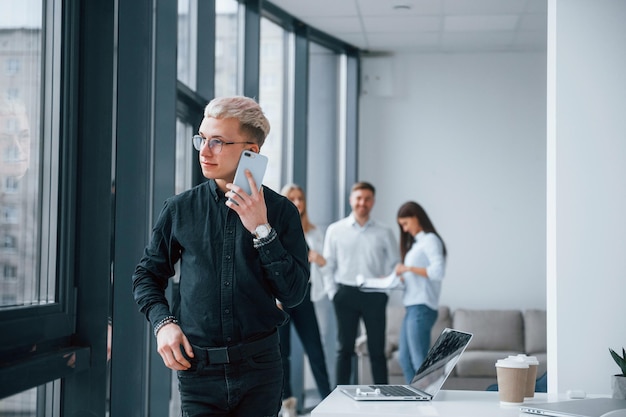 Junger Hipster mit Telefon steht vor einer Gruppe junger erfolgreicher Teams, die drinnen im Büro zusammenarbeiten und kommunizieren