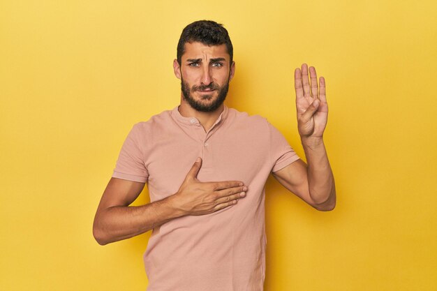 Junger hispanischer Mann auf gelbem Hintergrund legt einen Eid ab und legt die Hand auf die Brust