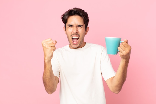 Junger hispanischer Mann, der aggressiv mit einem wütenden Ausdruck schreit und eine Kaffeetasse hält