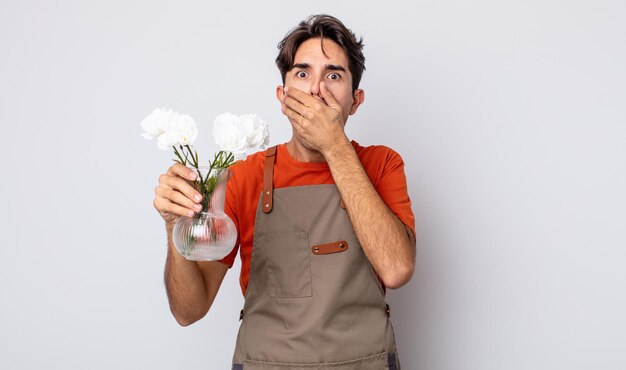 Junger hispanischer Mann, der den Mund mit den Händen mit einem schockierten bedeckt. Floristenkonzept