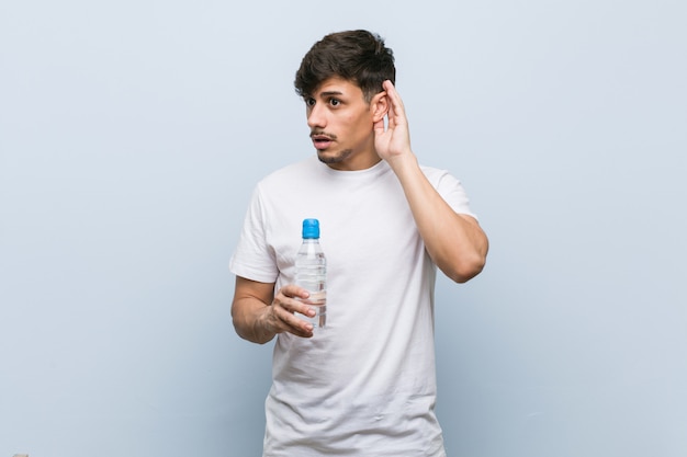 Junger hispanischer Mann, der eine Wasserflasche versucht, einen Klatsch zu hören hält.