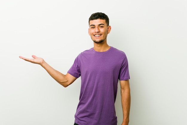 Junger hispanischer Mann, der einen Kopienraum auf einer Palme zeigt und eine andere Hand auf Taille hält