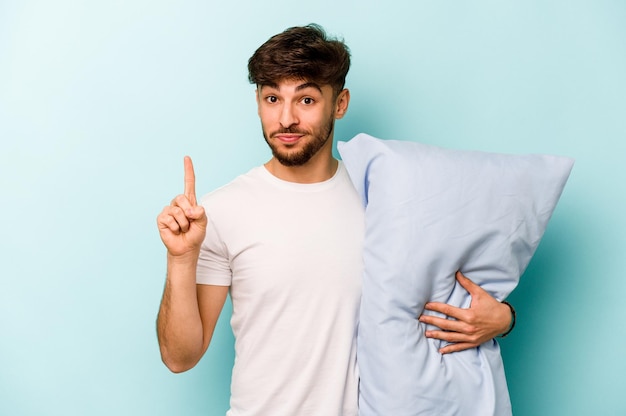 Junger hispanischer Mann, der einen Pyjama trägt und ein Kissen hält, das auf blauem Hintergrund isoliert ist und die Nummer eins mit dem Finger zeigt