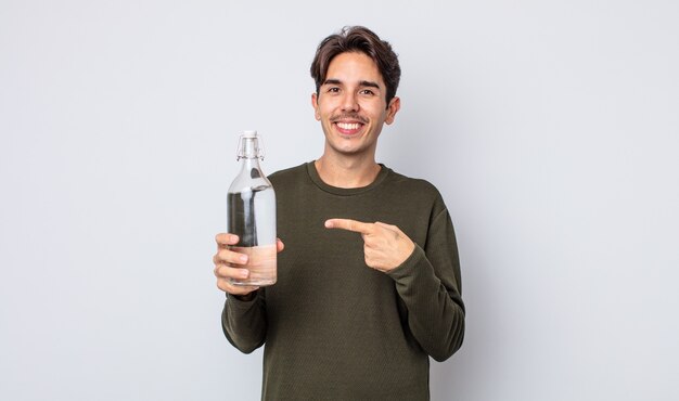 Junger hispanischer Mann, der fröhlich lächelt, sich glücklich fühlt und auf die Seite zeigt. Wasserflaschenkonzept