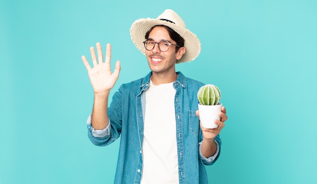 Junger hispanischer Mann, der glücklich lächelt, die Hand winkt, Sie begrüßt und begrüßt und einen Kaktus hält