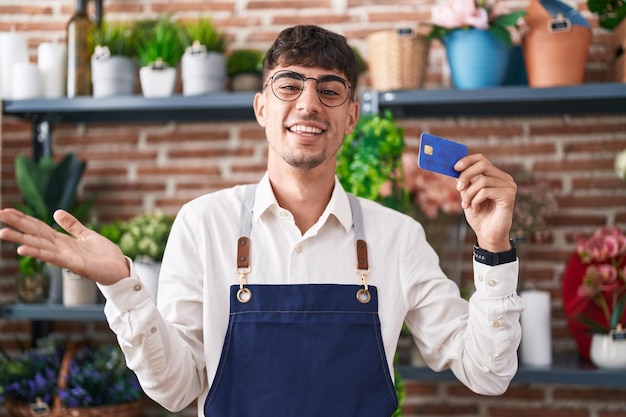 Junger hispanischer Mann, der in einem Blumengeschäft arbeitet und eine Kreditkarte in der Hand hält, feiert seinen Erfolg mit einem glücklichen Lächeln und einem Siegerausdruck mit erhobener Hand
