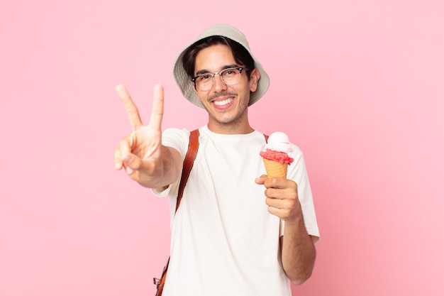 Junger hispanischer Mann, der lächelt und glücklich aussieht, Sieg oder Frieden gestikuliert und ein Eis hält