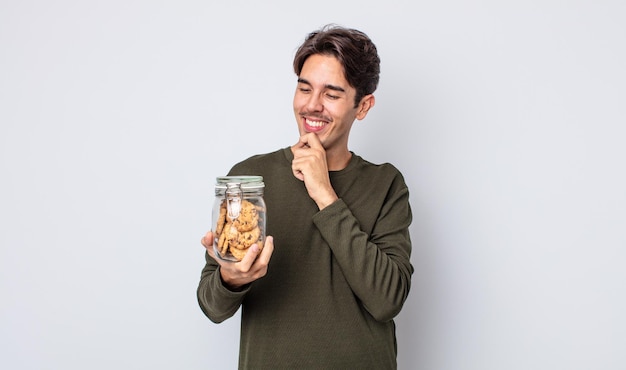 Junger hispanischer mann, der mit einem glücklichen, selbstbewussten ausdruck mit der hand am kinn lächelt. cookies-konzept