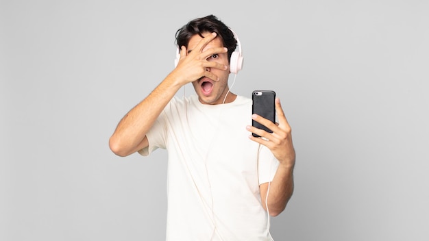 Junger hispanischer Mann, der schockiert, verängstigt oder verängstigt aussieht und das Gesicht mit der Hand mit Kopfhörern und Smartphone bedeckt