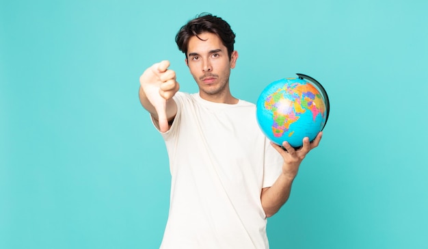 Junger hispanischer Mann, der sich mit dem Daumen nach unten zeigt und eine Weltkugelkarte hält