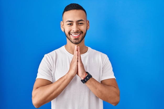 Junger hispanischer Mann, der über blauem Hintergrund steht und mit den Händen zusammen betet und um Vergebung bittet und selbstbewusst lächelt.