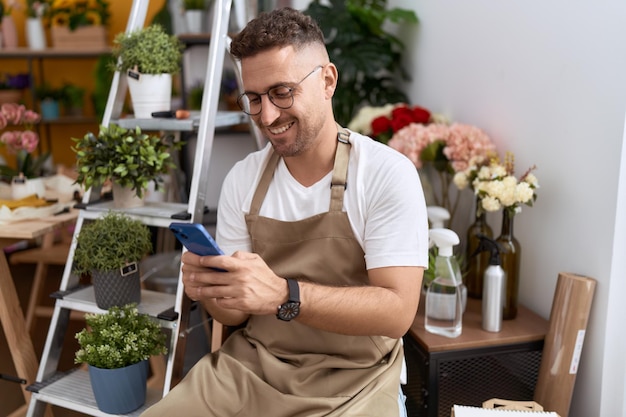 Junger hispanischer Mann Florist lächelt selbstbewusst mit Smartphone im Blumenladen