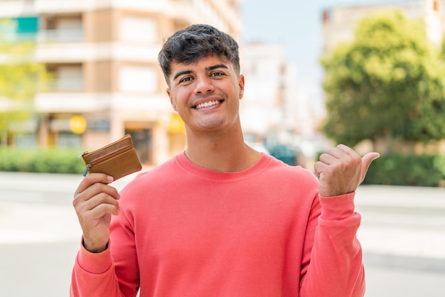 Junger hispanischer Mann hält eine Brieftasche im Freien und zeigt zur Seite, um ein Produkt zu präsentieren