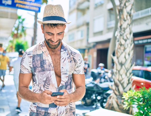 Junger hispanischer Mann im Urlaub lächelt glücklich mit Smartphone auf der Straße der Stadt