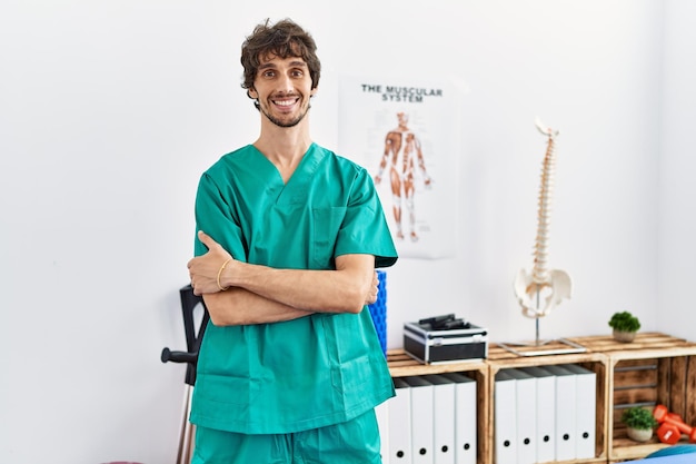 Junger hispanischer Mann in Physiotherapeutenuniform mit verschränkten Armen in der Physiotherapeutenklinik