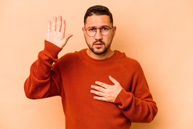 Junger hispanischer Mann isoliert auf beigem Hintergrund, der einen Eid ablegt und die Hand auf die Brust legt