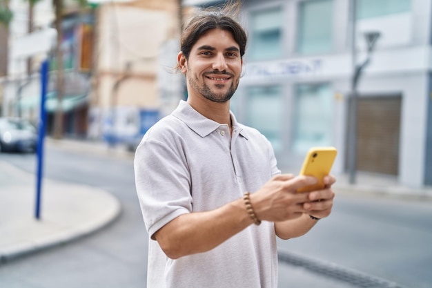 Junger hispanischer Mann lächelt selbstbewusst mit Smartphone auf der Straße