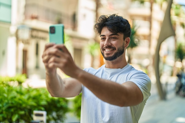 Junger hispanischer Mann lächelt selbstbewusst und macht Selfie mit dem Smartphone auf der Straße