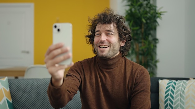 Junger hispanischer Mann macht Selfie per Smartphone, während er zu Hause auf dem Sofa sitzt