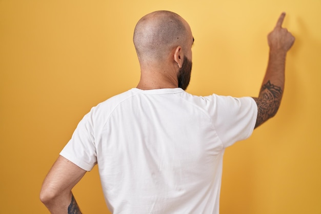 Junger hispanischer Mann mit Bart und Tätowierungen, der über gelbem Hintergrund steht und rückwärts zeigt und mit der Fingerhand nach vorne zeigt