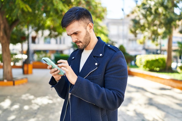 Junger hispanischer Mann mit Smartphone mit ernster Miene im Park