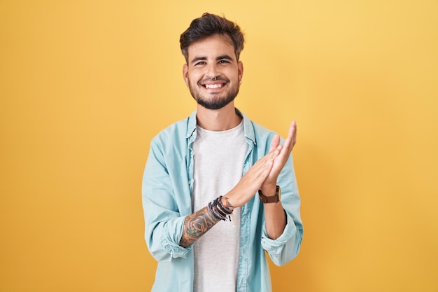 Junger hispanischer Mann mit Tätowierungen, der vor gelbem Hintergrund steht und klatscht und applaudiert, glücklich und freudig lächelnd, stolze Hände zusammen