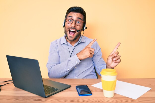 Junger hispanischer Mann sitzt am Schreibtisch und trägt ein Vermittlungs-Headset im Callcenter-Büro, lächelt und blickt in die Kamera, während er mit zwei Händen und Fingern zur Seite zeigt