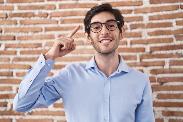 Junger hispanischer Mann steht vor dem Hintergrund einer Backsteinmauer und zeigt lächelnd mit einem Finger auf den Kopf. Tolle Idee oder guter Gedanke, gute Erinnerung