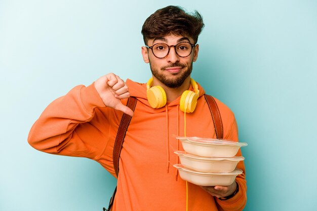 Junger hispanischer Student, der eine Tupperware hält, die auf blauem Hintergrund isoliert ist und eine Abneigungsgeste mit dem Daumen nach unten zeigt