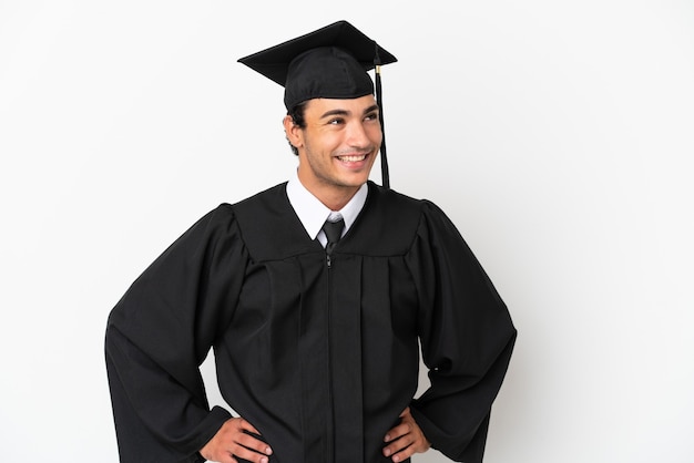 Junger Hochschulabsolvent über isoliertem weißem Hintergrund posiert mit Armen an der Hüfte und lächelt
