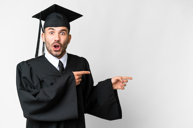 Junger Hochschulabsolvent über isoliertem weißem Hintergrund überrascht und zeigt zur Seite