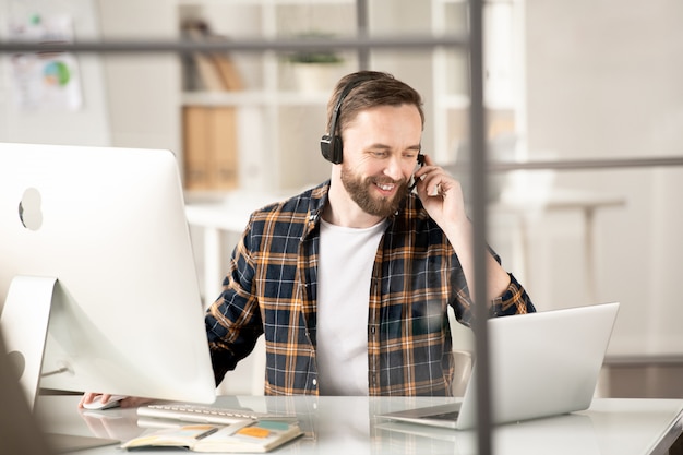 Junger Hotlineberater mit Headset, der vor Laptop und Computer sitzt und mit einem der Kunden online spricht