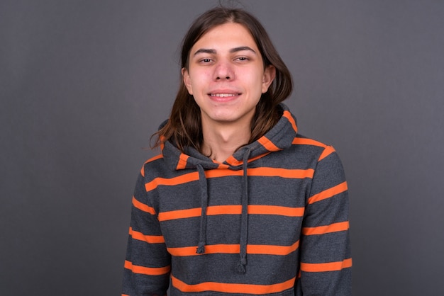 Foto junger hübscher androgyner mann mit langen haaren, der kapuzenpulli gegen graue wand trägt