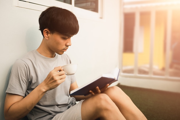 Junger hübscher asiatischer Mann, der Buch liest und Tee auf Raumbalkon in der Freizeit vom Arbeiten zu Hause, Wissens- und Lernkonzept trinkt