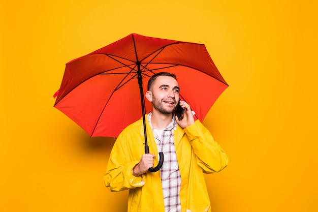 Junger hübscher bärtiger Mann im gelben Regenmantel mit rotem Regenschirm, der durch Handy lokalisiert über orange Hintergrund spricht