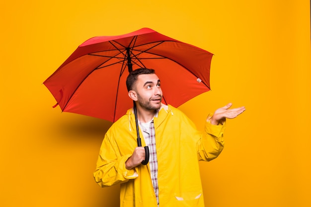 Junger hübscher bärtiger Mann im gelben Regenmantel mit rotem Regenschirm, der versucht, zu sehen, ob sein Regen über orange Hintergrund isoliert