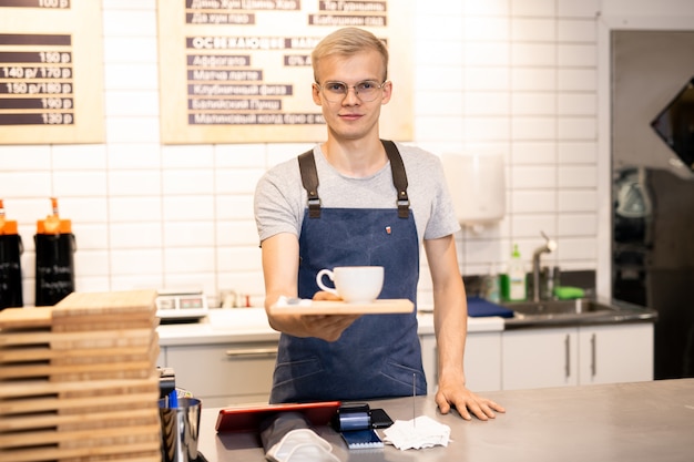 Junger hübscher blonder Kellner in Brille und Arbeitskleidung, die Ihnen Tablett mit Tasse Kaffee geben, während Sie an der Theke stehen