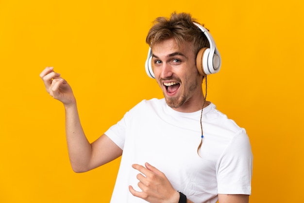 Junger hübscher blonder Mann lokalisiert auf gelbem Hintergrund, der Musik hört und Gitarrengeste tut