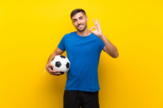 Junger hübscher Fußballspielermann über der lokalisierten gelben Wand, die okayzeichen mit den Fingern zeigt