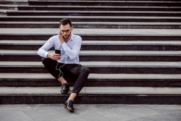 Junger hübscher geeky Mann elegant gekleidet, der auf der Treppe draußen sitzt und Musik auf Smartphone hört.