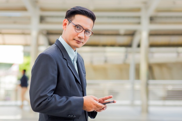 Junger hübscher Geschäftsmann Asiens mit seinem Smartphone, der auf Gehweg der modernen Stadt steht.